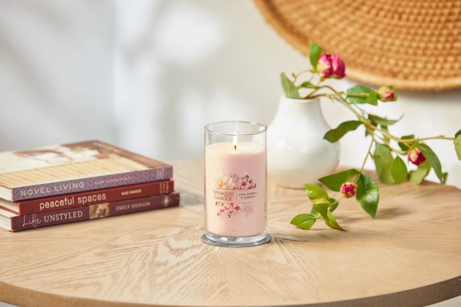 lit pink cherry and vanilla signature medium pillar candle on wooden table next to books and a flower vase