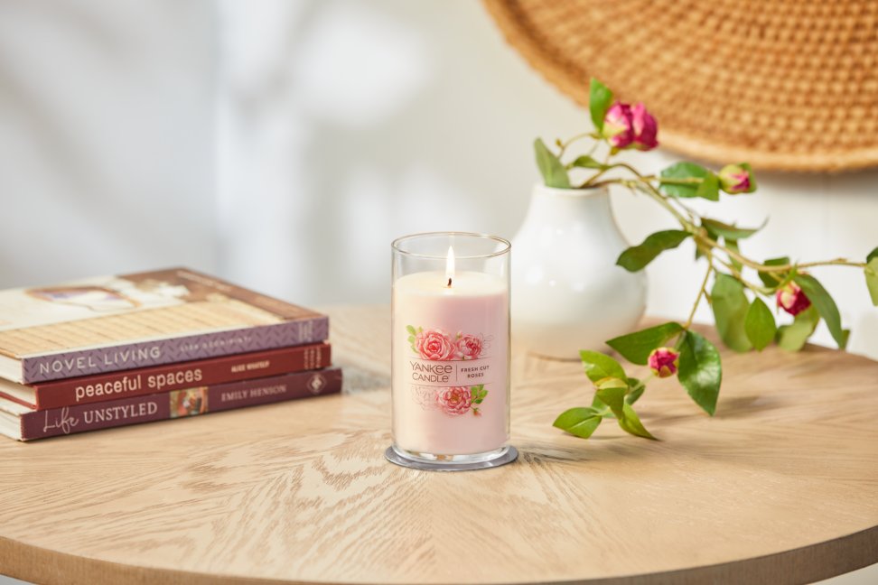 lit fresh cut roses signature medium pillar candle on wooden table next to books and a flower vase