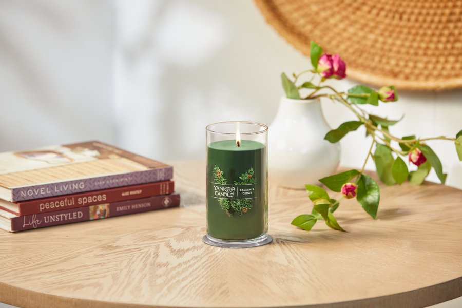 lit balsam and cedar signature medium pillar candle on wooden table next to books and a flower vase