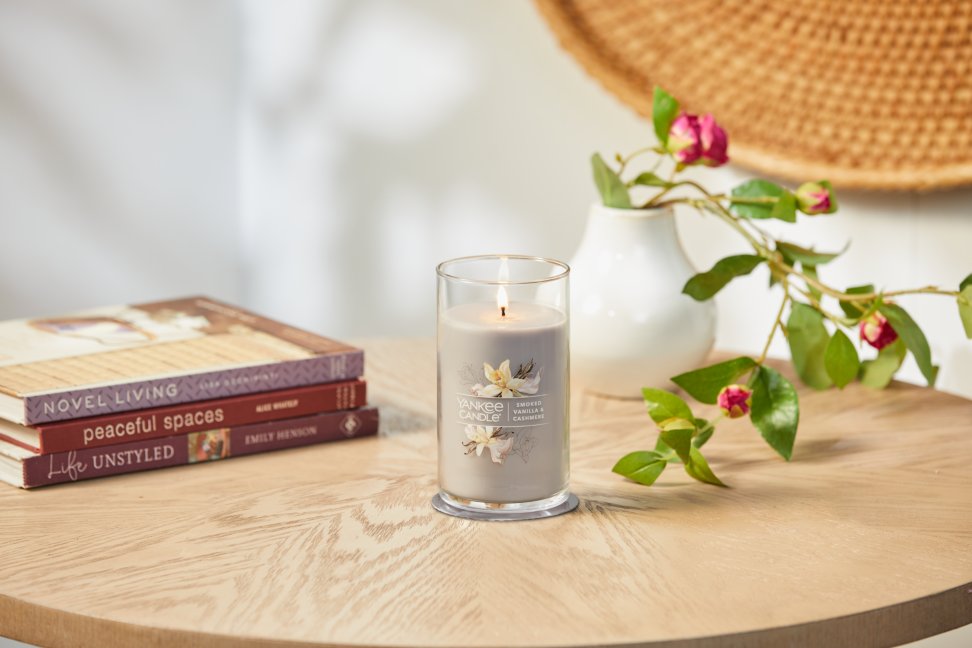 lit smoked vanilla and cashmere signature medium pillar candle on wooden table next to books and a flower vase