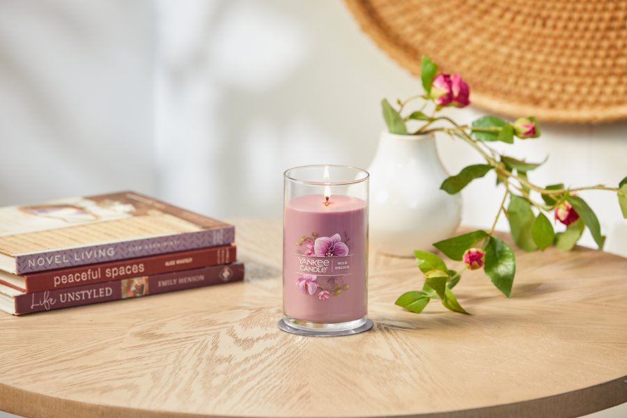 lit wild orchid signature medium pillar candle on wooden table next to books and a flower vase