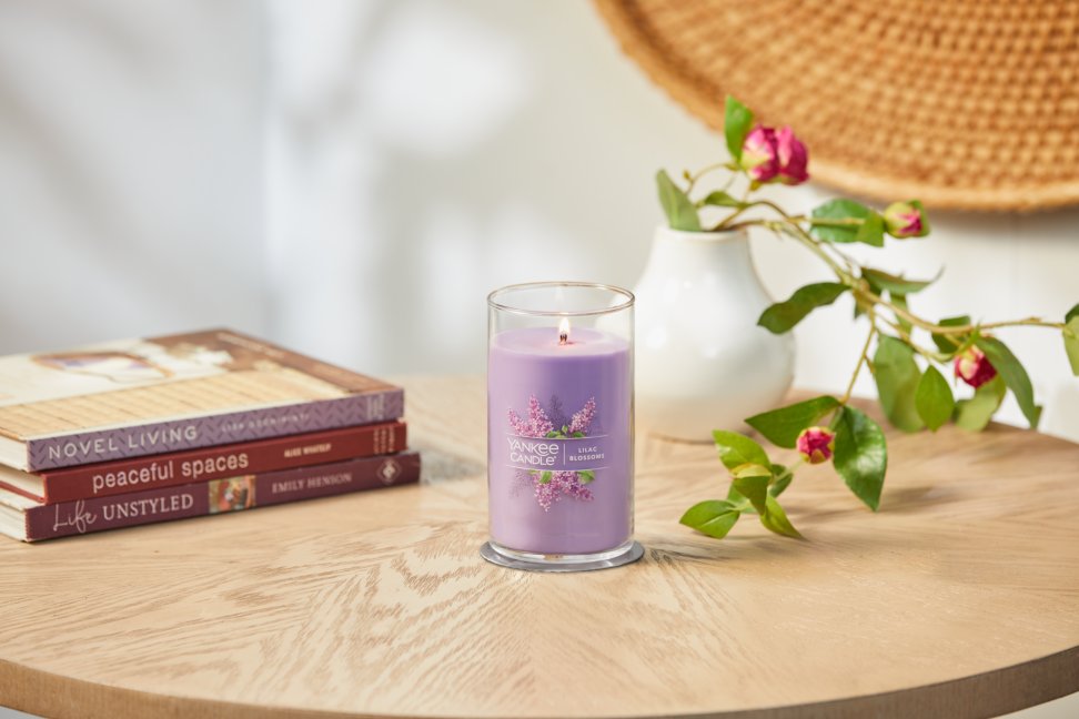 lit lilac blossoms signature medium pillar candle on wooden table next to books and roses
