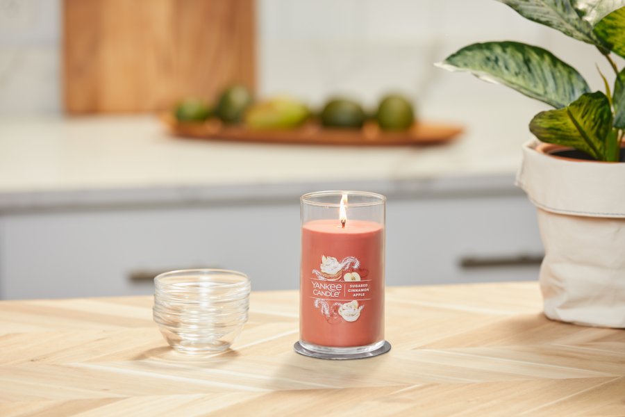 lit sugared cinnamon apple signature medium pillar candle on wooden counter next to a potted plant