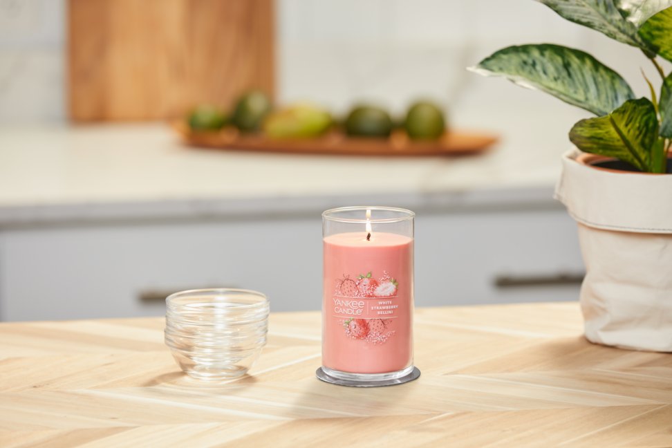 lit white strawberry bellini signature medium pillar candle on wooden counter next to a potted plant