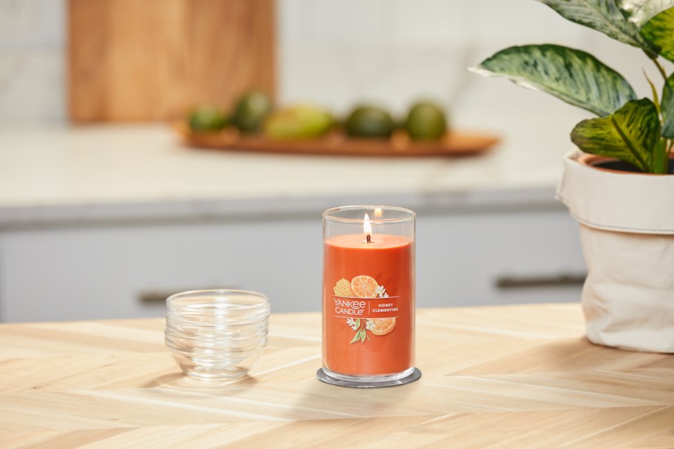lit honey clementine signature medium pillar candle on wooden counter next to a potted plant