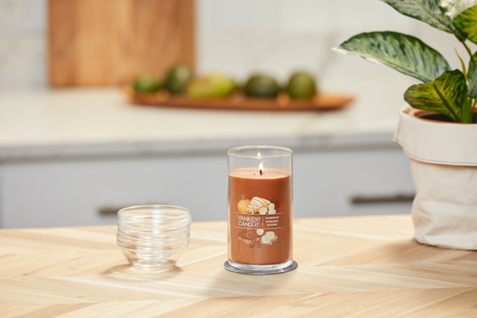 lit pumpkin banana scone signature medium pillar candle on wooden counter next to a potted plant