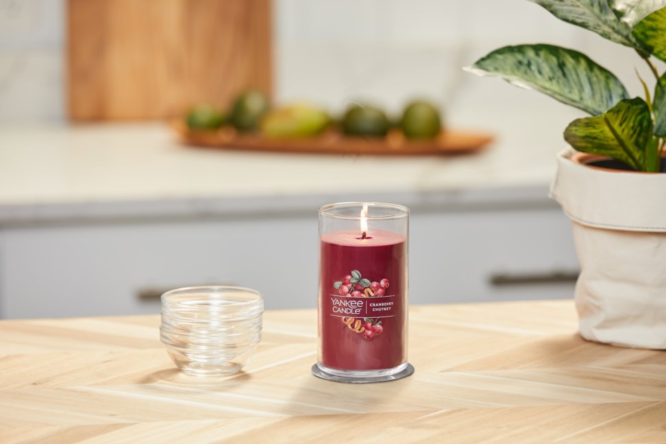 lit cranberry chutney signature medium pillar candle on wooden counter next to a potted plant
