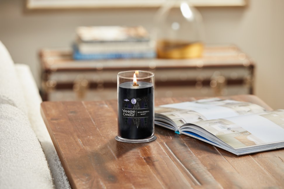 lit midsummers night signature medium pillar candle on wooden table next to a book