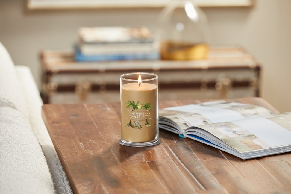 lit sun and sand signature medium pillar candle on wooden table next to a book