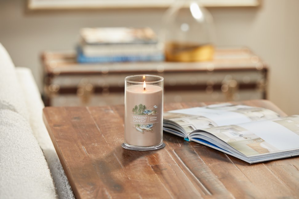 lit seaside woods signature medium pillar candle on wooden table next to a book