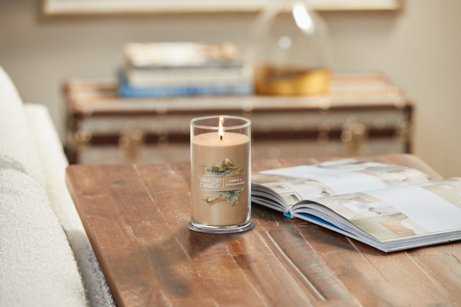lit amber and sandalwood signature medium pillar candle on wooden table next to open book