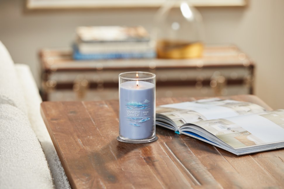 lit ocean air signature medium pillar candle on wooden table next to a book