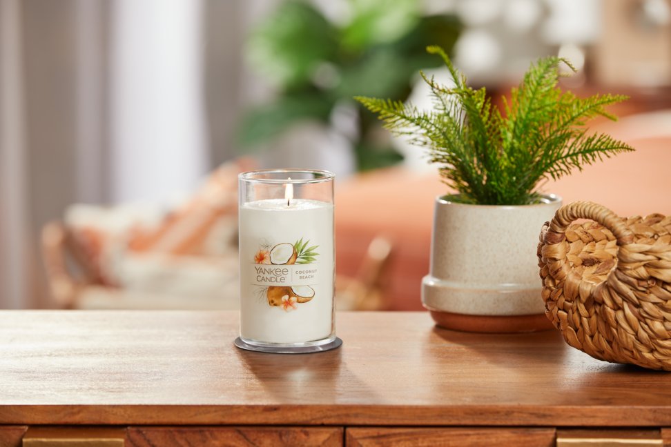 lit coconut beach signature medium pillar candle on wooden table next to a potted plant and basket
