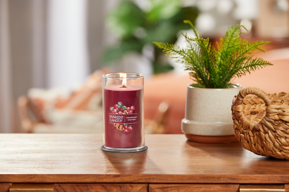 lit cranberry chutney signature medium pillar candle on wooden table next to a potted plant and basket