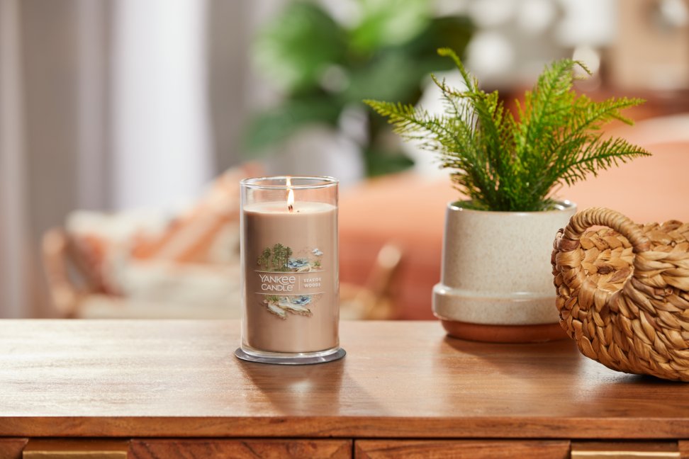 lit seaside woods signature medium pillar candle on wooden table next to a potted plant and basket