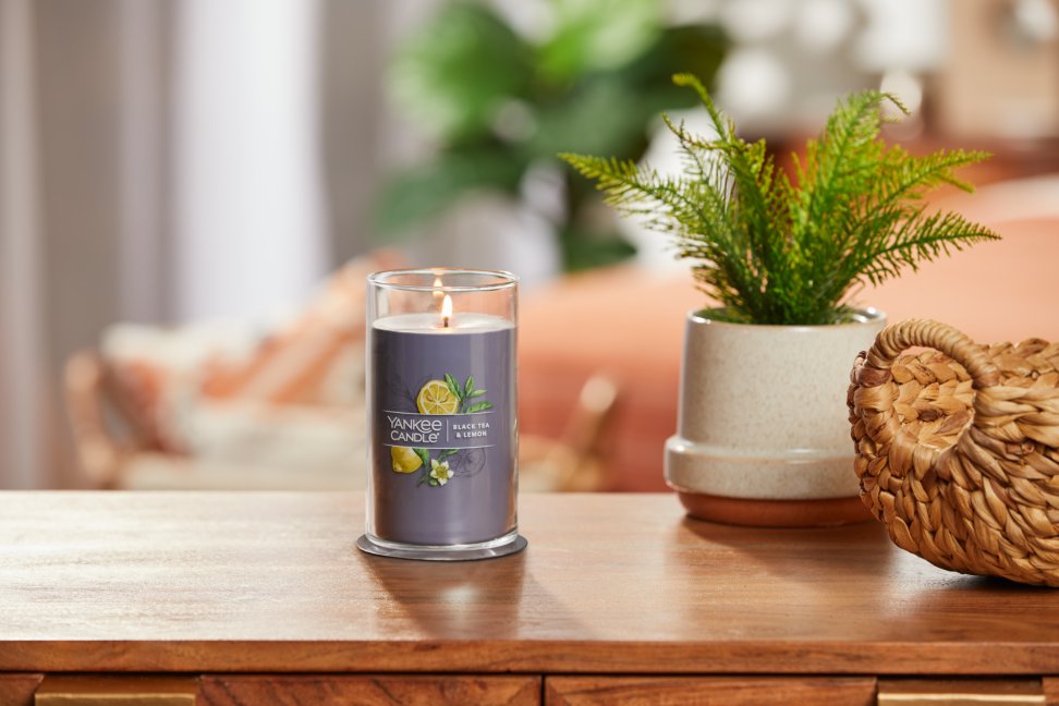 lit black tea and lemon signature medium pillar candle on wooden table next to a potted plant and basket