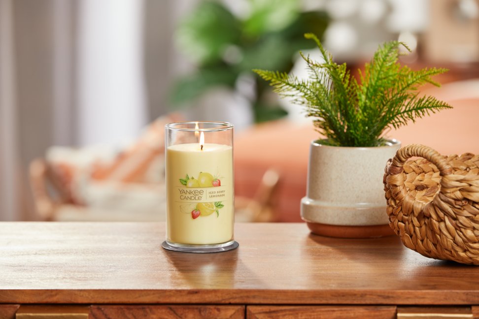 lit iced berry lemonade signature medium pillar candle on wooden table next to a potted plant and basket