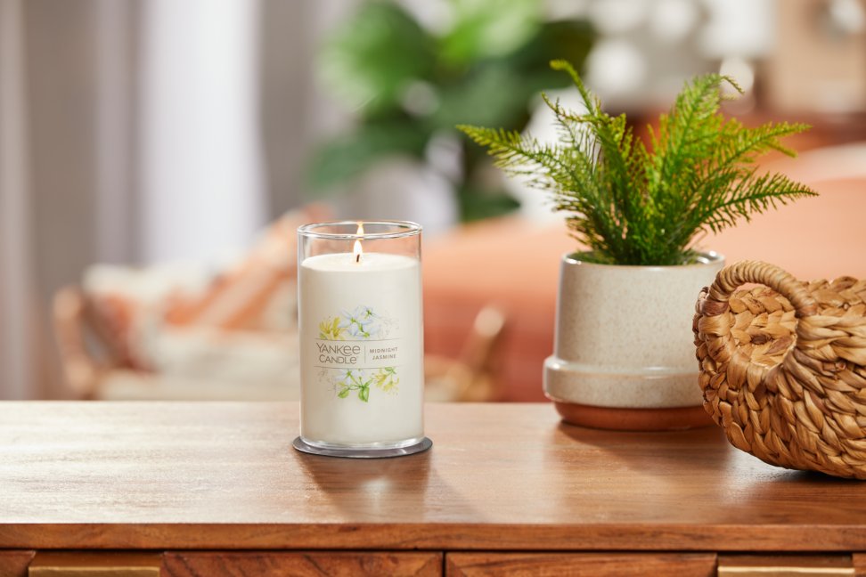 lit midnight jasmine signature medium pillar candle on wooden table next to a potted plant and basket