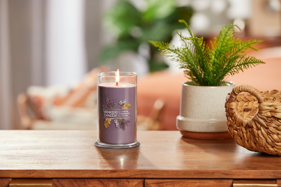 lit dried lavender and oak signature medium pillar candle on wooden table next to a potted plant and basket