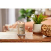 lit amber and sandalwood signature medium pillar candle on wooden table next to a potted plant and basket image number 3