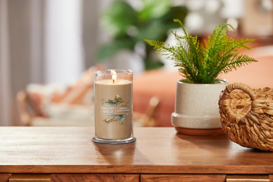 lit amber and sandalwood signature medium pillar candle on wooden table next to a potted plant and basket