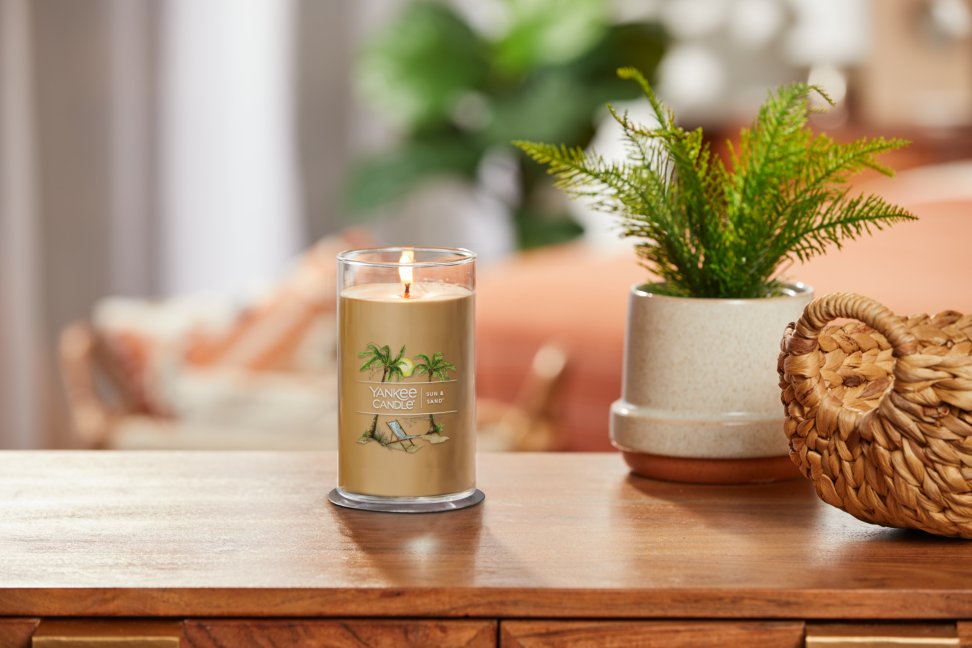 lit sun and sand signature medium pillar candle on wooden table next to a potted plant and basket