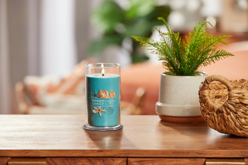 lit catching rays signature medium pillar candle on wooden table next to a potted plant and basket
