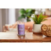 lit lilac blossoms signature medium pillar candle on wooden table next to a potted plant and basket image number 3