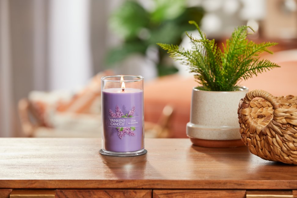 lit lilac blossoms signature medium pillar candle on wooden table next to a potted plant and basket