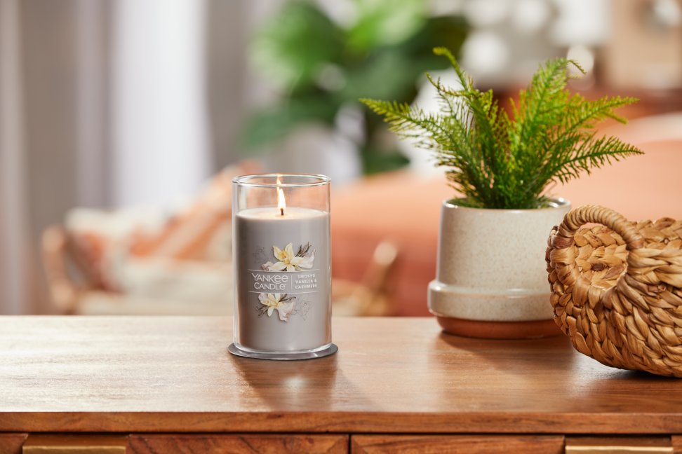 lit smoked vanilla and cashmere signature medium pillar candle on wooden table next to a potted plant and basket