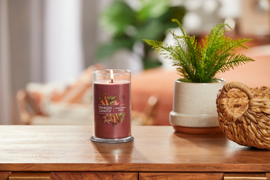 lit home sweet home signature medium pillar candle on wooden table next to a potted plant and basket