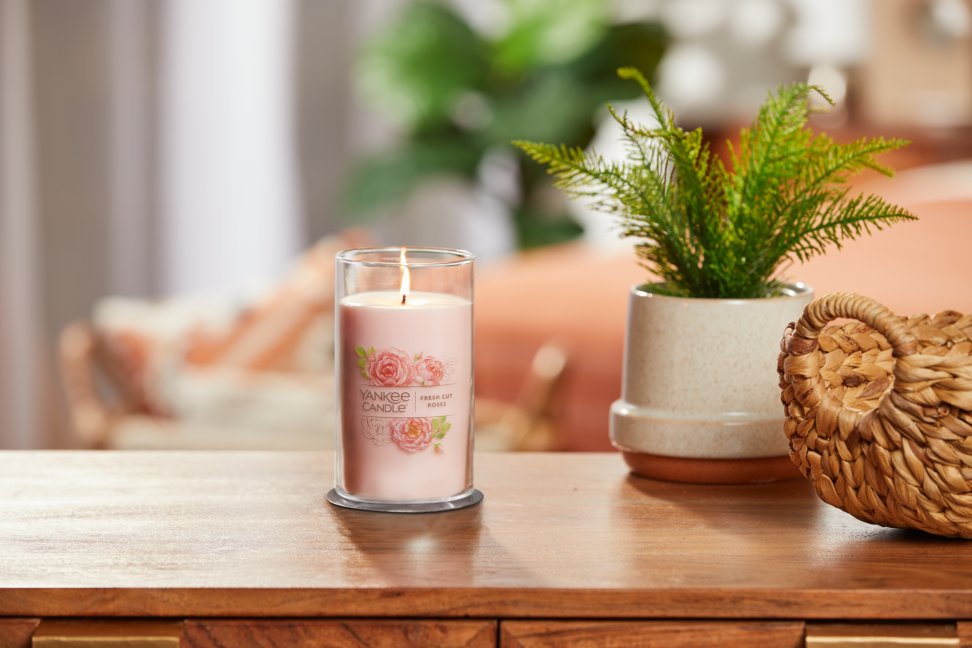 lit fresh cut roses signature medium pillar candle on wooden table next to a potted plant and basket