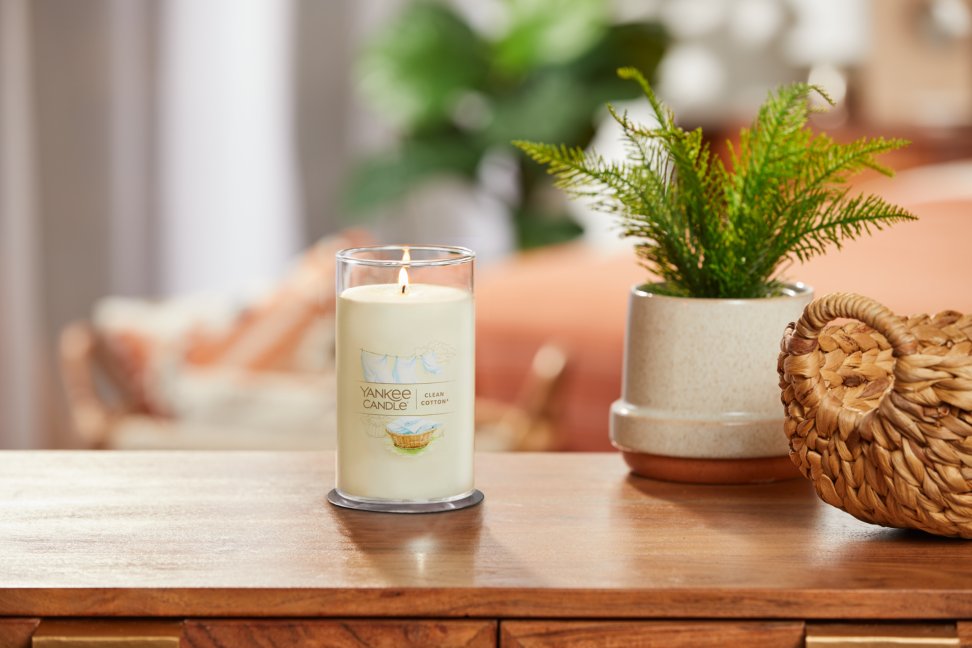 lit clean cotton signature medium pillar candle on wooden table next to a potted plant and basket