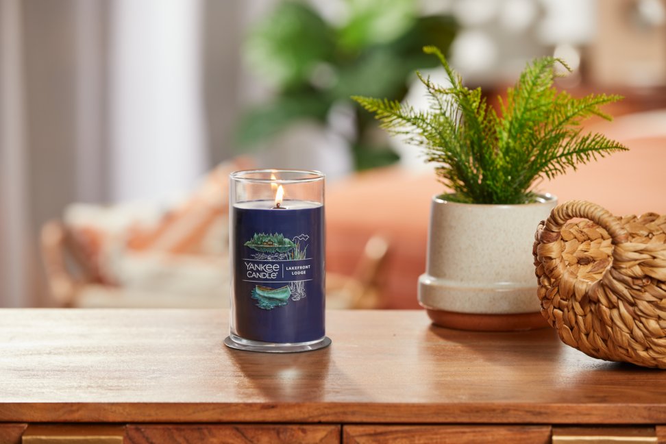 lit lakefront lodge signature medium pillar candle on wooden table next to a potted plant and basket