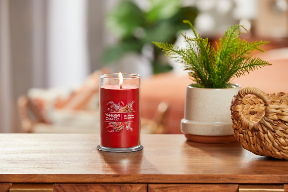 lit sparkling cinnamon signature medium pillar candle on wooden table next to a potted plant and basket