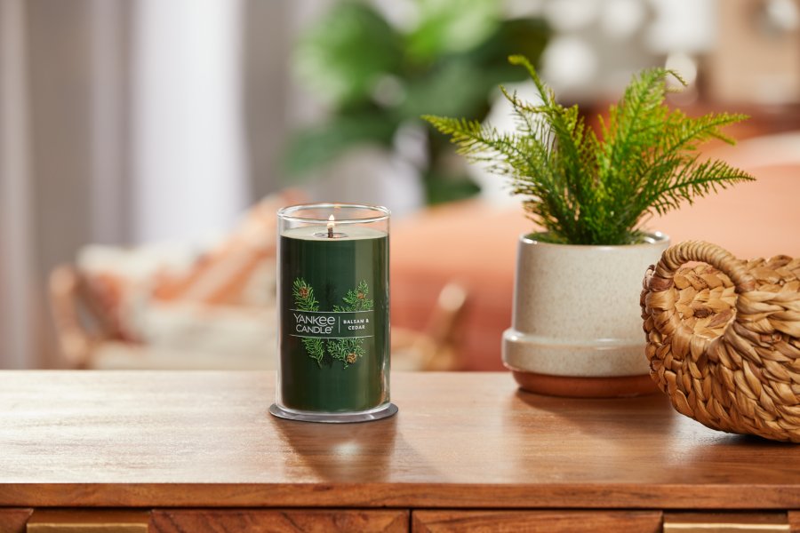 lit balsam and cedar signature medium pillar candle on wooden table next to a potted plant and basket