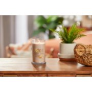 lit vanilla creme brulee signature medium pillar candle on wooden table next to a potted plant and basket image number 3