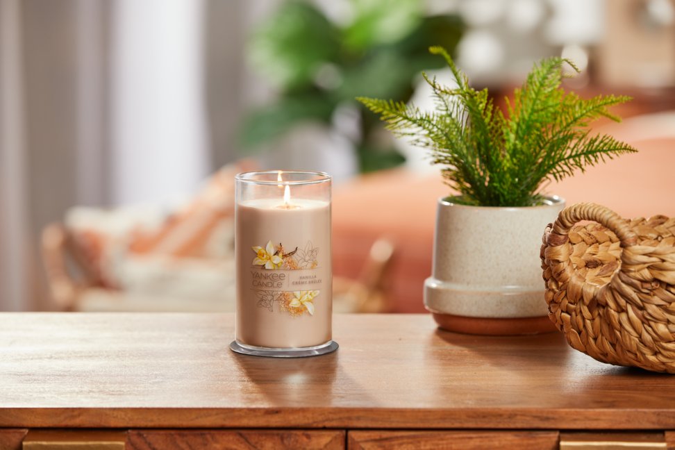 lit vanilla creme brulee signature medium pillar candle on wooden table next to a potted plant and basket