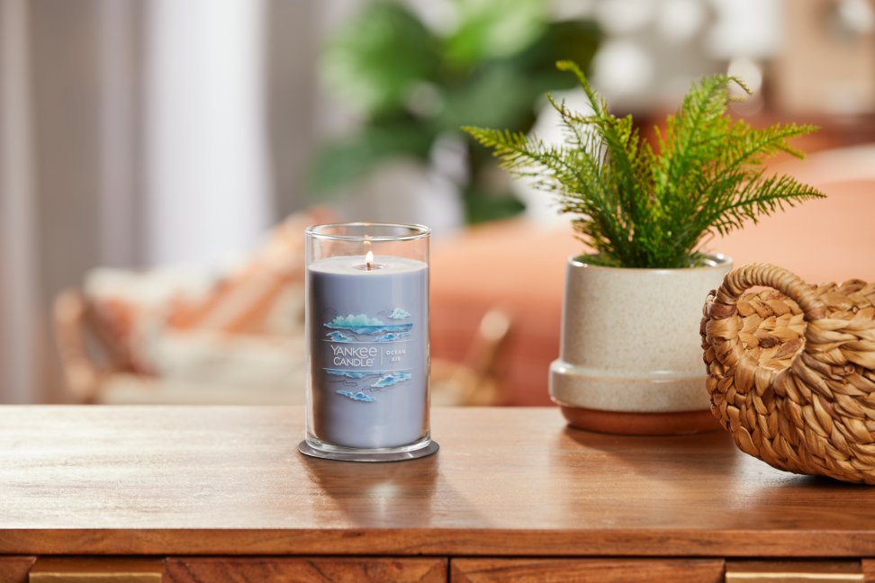 lit ocean air signature medium pillar candle on wooden table next to a potted plant and basket