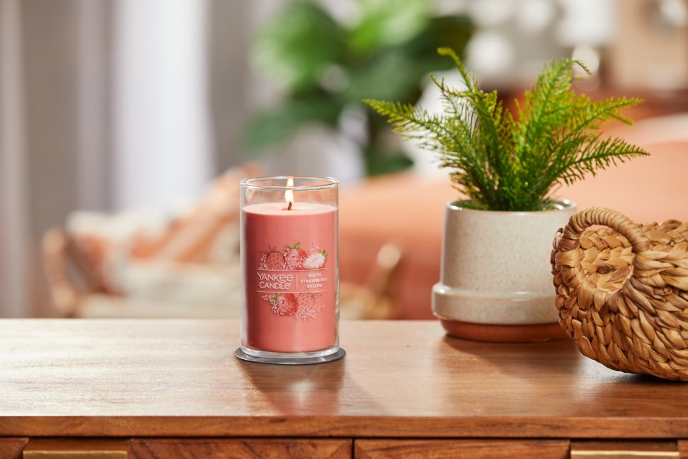 lit white strawberry bellini signature medium pillar candle on wooden table next to a potted plant and basket