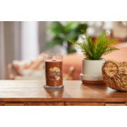 lit pumpkin banana scone signature medium pillar candle on wooden table next to a potted plant and basket image number 3