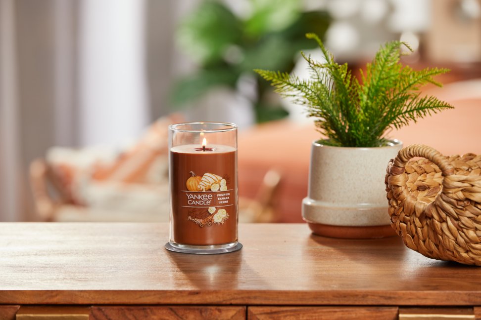 lit pumpkin banana scone signature medium pillar candle on wooden table next to a potted plant and basket