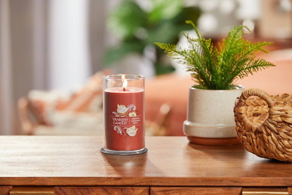 lit sugared cinnamon apple signature medium pillar candle on wooden table next to a potted plant and basket