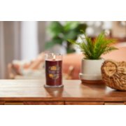 lit holiday zest signature medium pillar candle on wooden table next to a potted plant and basket image number 3