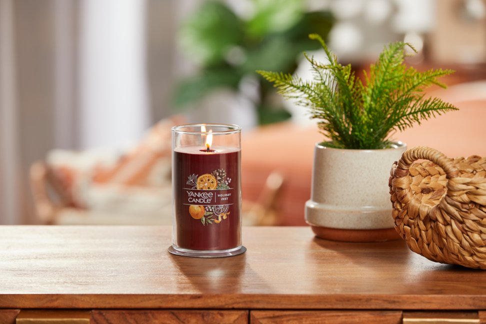 lit holiday zest signature medium pillar candle on wooden table next to a potted plant and basket