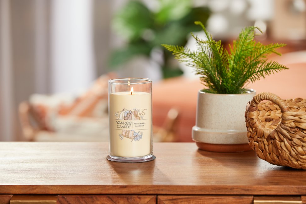 lit soft wool and amber signature medium pillar candle on wooden table next to a potted plant and basket