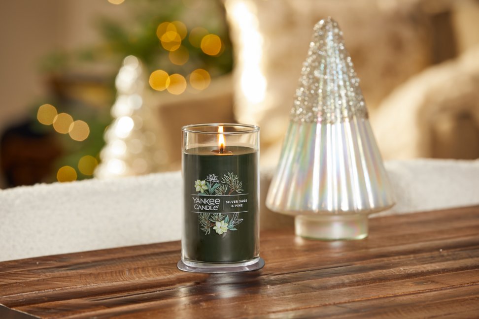 lit silver sage and pine signature medium pillar candle on wooden table next to a white and silver glitter tree