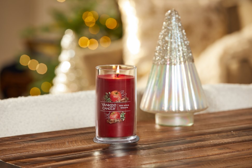 lit red apple wreath signature medium pillar candle on wooden table next to a white and silver glitter tree