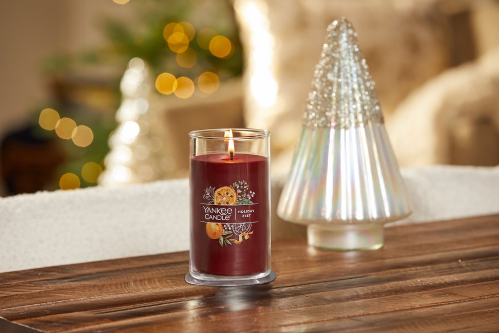 lit holiday zest signature medium pillar candle on wooden table next to a white and silver glitter tree