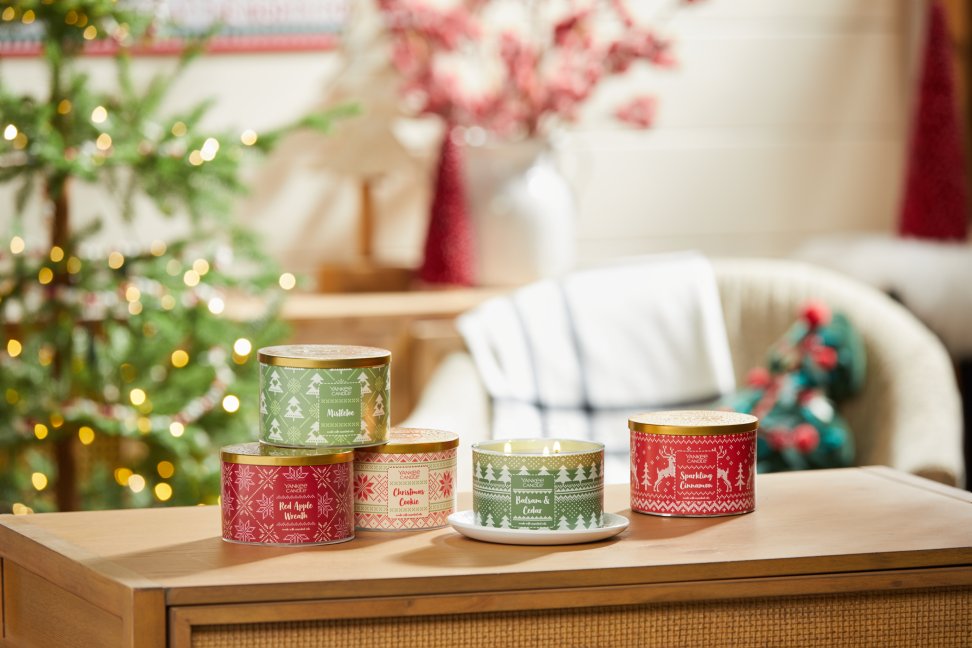 the holiday knits collection mistletoe 3 wick candle, red apple wreath 3 wick candle, balsam and cedar 3 wick candle, and sparkling cinnamon 3 wick candle on a wood table in a living room setting